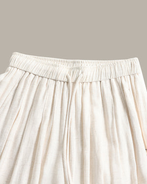 Close-up of a pants in a light cream or off-white fabric, showing the gathered elastic waistband and hanging drawstring ties against a solid taupe background.