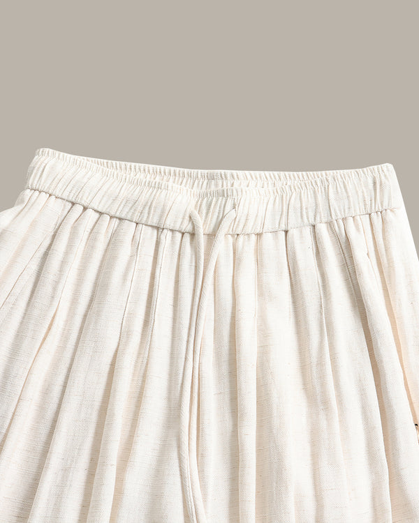Close-up of a pants in a light cream or off-white fabric, showing the gathered elastic waistband and hanging drawstring ties against a solid taupe background.