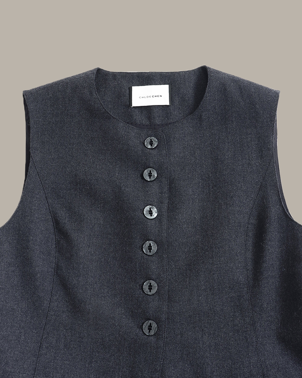A dark gray sleeveless vest with a button-down front and two front pockets, displayed flat against a solid taupe background.