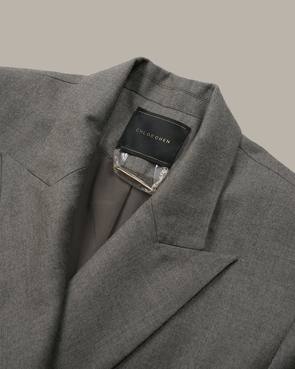 Close-up of a gray textured blazer jacket with a black brand label reading CHLOE GREN on the collar, shown against a solid taupe background.