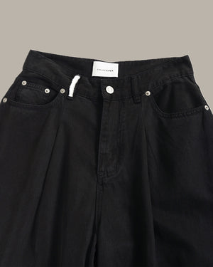 Close-up of black denim pants showing the waistband, front pockets, rivets, and a white drawstring against a plain taupe background.