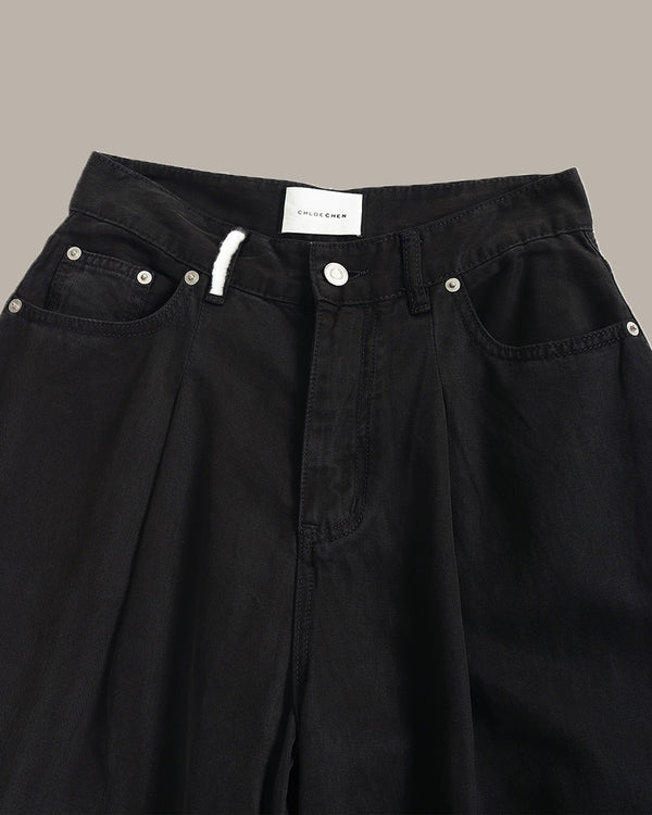 Close-up of black denim pants showing the waistband, front pockets, rivets, and a white drawstring against a plain taupe background.