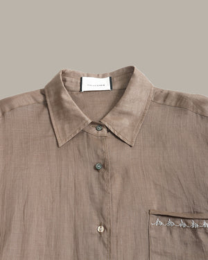 A close-up of a taupe linen button-down shirt with a pointed collar, button placket, and a chest pocket featuring embroidered details, set against a solid taupe background.