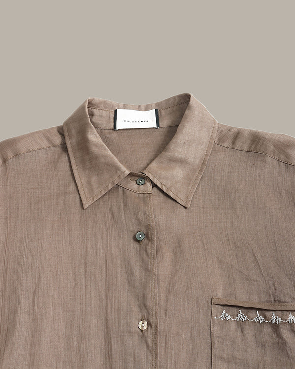 A close-up of a taupe linen button-down shirt with a pointed collar, button placket, and a chest pocket featuring embroidered details, set against a solid taupe background.
