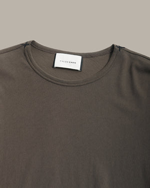 Close-up of a dark brown crew-neck t-shirt by Chloe Chew, showing the white brand label at the collar against a solid taupe background.