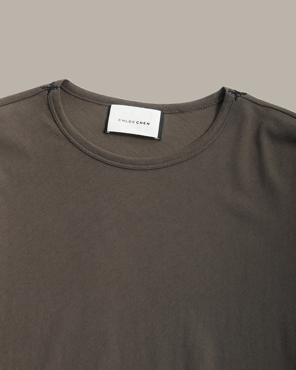 Close-up of a dark brown crew-neck t-shirt by Chloe Chew, showing the white brand label at the collar against a solid taupe background.