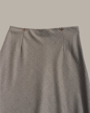 Close-up of a gray knit skirt with vertical center pleats and small brown embroidered details near the waistband, shown against a solid taupe background.
