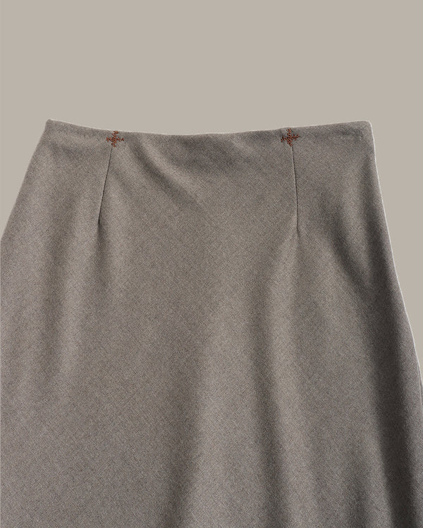 Close-up of a gray knit skirt with vertical center pleats and small brown embroidered details near the waistband, shown against a solid taupe background.