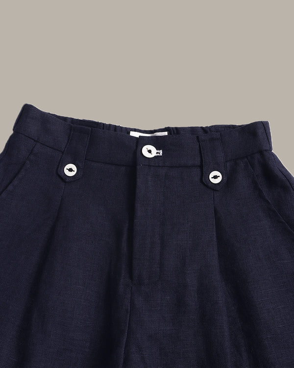 Close-up of dark navy pleated trousers showing the waistband, button details, and textured fabric against a solid taupe background.