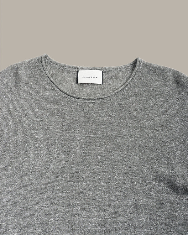Close-up of a gray knit crewneck sweater with a white neck label reading 'SILK ROCKS', shown against a solid taupe background.