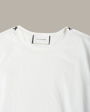 A close-up of a white crewneck t-shirt by Chloé Chloé, featuring a white neck label with black text and small black butterfly details at the shoulder seams, set against a solid taupe background.