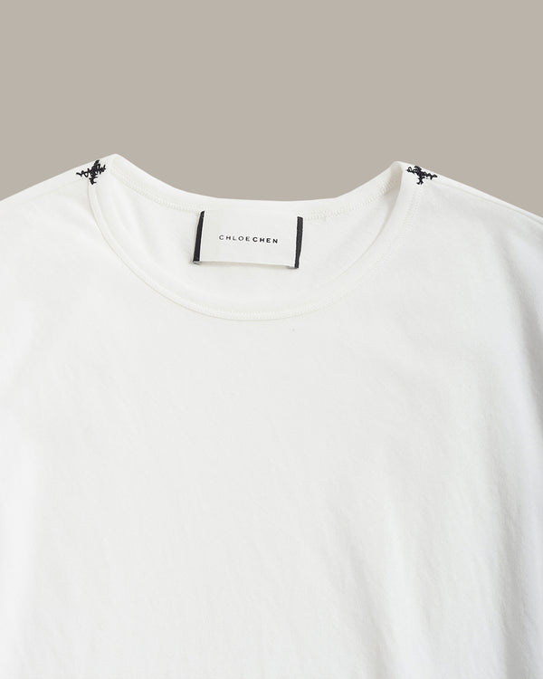 A close-up of a white crewneck t-shirt by Chloé Chloé, featuring a white neck label with black text and small black butterfly details at the shoulder seams, set against a solid taupe background.
