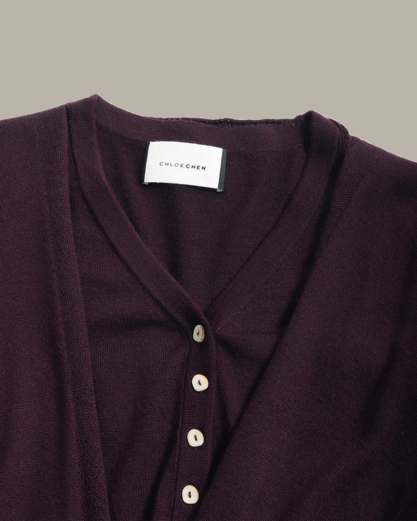 Close-up of a dark plum purple knit cardigan by Chloé Gémen, featuring a V-neckline and cream-colored buttons, with a white brand label visible at the collar.