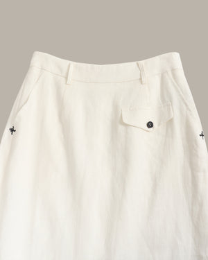 Close-up of an off-white linen skirt showing the waistband, belt loops, a buttoned flap pocket, and small embroidered cross details on the sides.