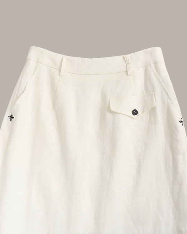Close-up of an off-white linen skirt showing the waistband, belt loops, a buttoned flap pocket, and small embroidered cross details on the sides.