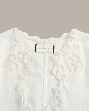 Close-up of a white embroidered blouse with floral eyelet lace detailing and a visible CHLOE CHEN neck label, set against a solid taupe background.