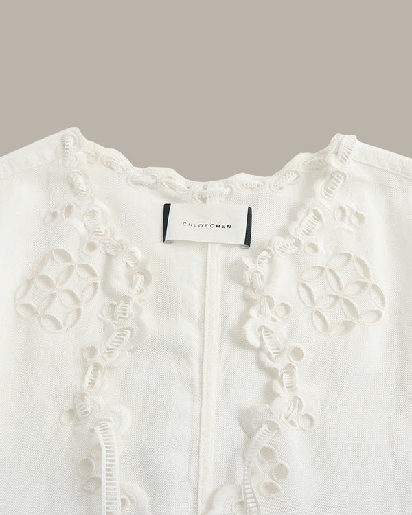 Close-up of a white embroidered blouse with floral eyelet lace detailing and a visible CHLOE CHEN neck label, set against a solid taupe background.