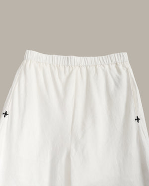 White wide-leg trousers with an elastic waistband and small black embroidered cross motifs on the sides, shown against a solid taupe background.