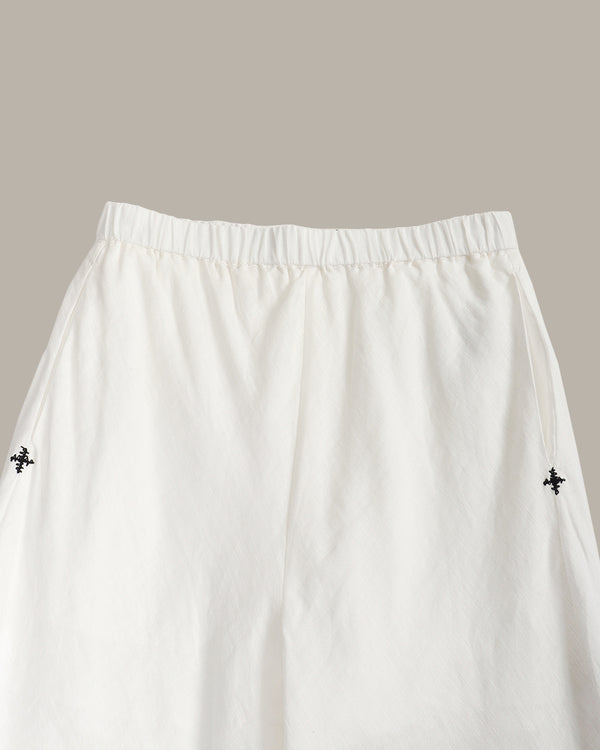 White wide-leg trousers with an elastic waistband and small black embroidered cross motifs on the sides, shown against a solid taupe background.