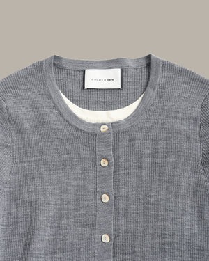Close-up of a gray ribbed knit henley shirt by Colorener, featuring a buttoned placket and a white inner collar lining, set against a solid taupe background.