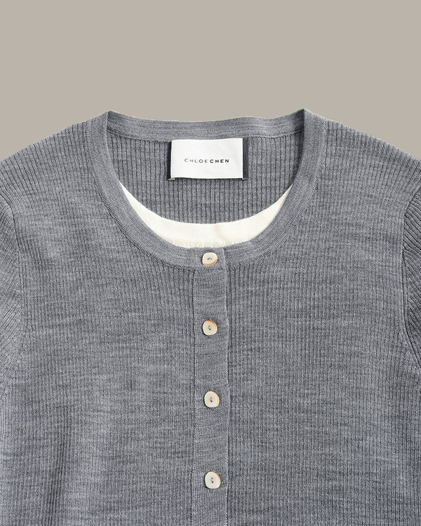 Close-up of a gray ribbed knit henley shirt by Colorener, featuring a buttoned placket and a white inner collar lining, set against a solid taupe background.