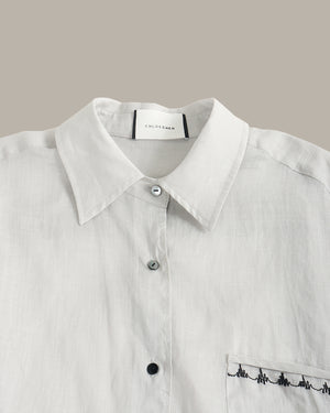Close-up of a white button-up shirt with a pointed collar, silver buttons, and black embroidered detailing on the chest pocket, shown against a solid taupe background.