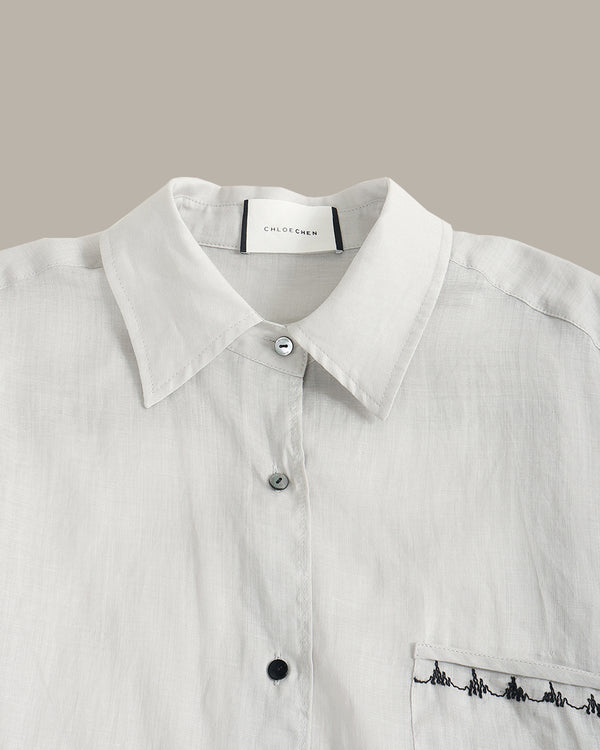 Close-up of a white button-up shirt with a pointed collar, silver buttons, and black embroidered detailing on the chest pocket, shown against a solid taupe background.