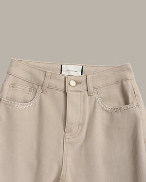 Close-up of a pair of light beige denim jeans featuring decorative braided stitching along the pocket edges and a white waistband label.