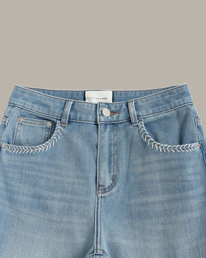 Close-up of a pair of light blue denim jeans showing the front waistband, button closure, belt loops, and decorative white contrast stitching around the front pockets. A white brand label reading 'MURDER' is visible at the center waistband.