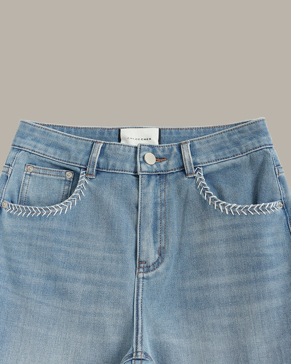 Close-up of a pair of light blue denim jeans showing the front waistband, button closure, belt loops, and decorative white contrast stitching around the front pockets. A white brand label reading 'MURDER' is visible at the center waistband.