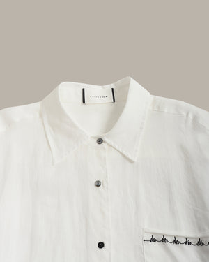 Close-up of a white collared button-up shirt with dark buttons and black embroidered line detail along the pocket edge, shown against a plain warm gray background.