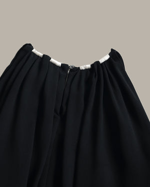 Close-up of a black pleated top with gathered elastic waistband and white trim details, shown against a solid taupe background.