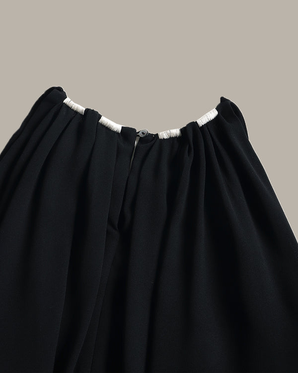 Close-up of a black pleated top with gathered elastic waistband and white trim details, shown against a solid taupe background.