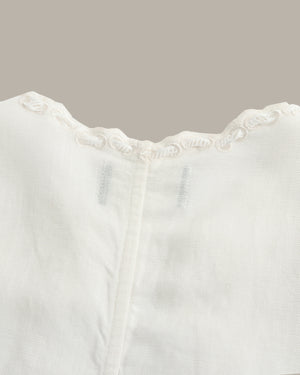 Close-up of a white textured fabric garment with a scalloped embroidered neckline and a central vertical seam, shown against a solid taupe background.