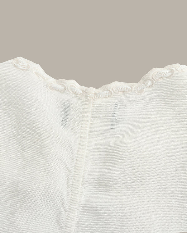 Close-up of a white textured fabric garment with a scalloped embroidered neckline and a central vertical seam, shown against a solid taupe background.