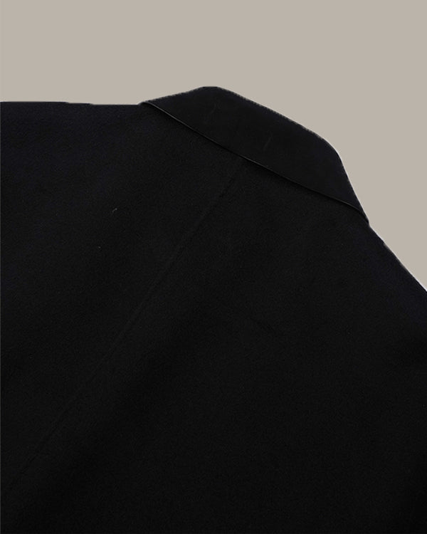 Close-up of a black garment, showing the shoulder and upper chest area with a subtle texture, set against a solid taupe background.
