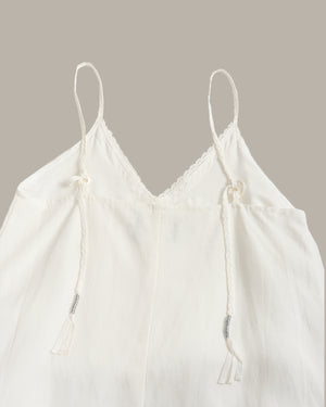An off-white cotton camisole top with thin adjustable drawstring spaghetti straps, a scalloped lace V-neckline, and a relaxed silhouette, shown against a solid taupe background.