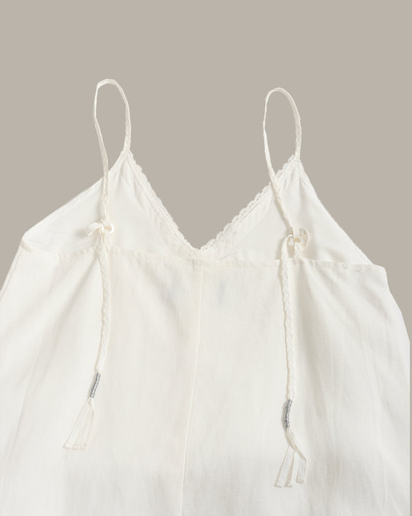 An off-white cotton camisole top with thin adjustable drawstring spaghetti straps, a scalloped lace V-neckline, and a relaxed silhouette, shown against a solid taupe background.