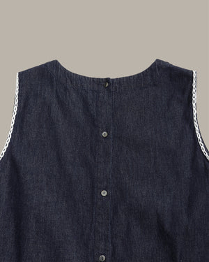 Close-up of a sleeveless dark indigo denim top with a button-down front and white chain trim along the shoulders, shown against a solid taupe background.
