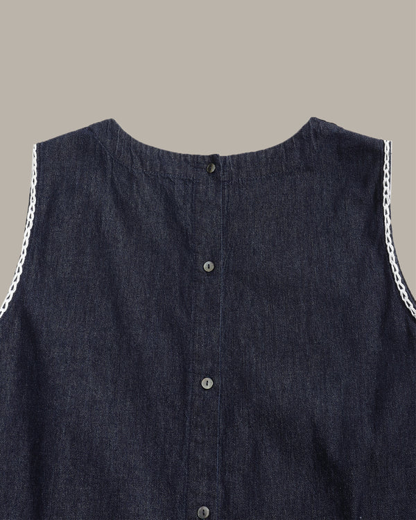 Close-up of a sleeveless dark indigo denim top with a button-down front and white chain trim along the shoulders, shown against a solid taupe background.