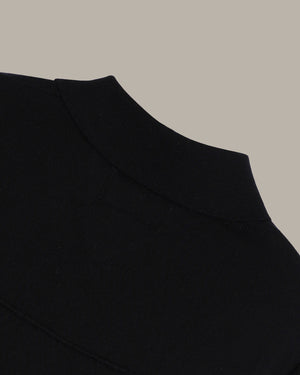 Close-up detail shot of a black knit garment, showing the collar and shoulder area against a solid taupe background.
