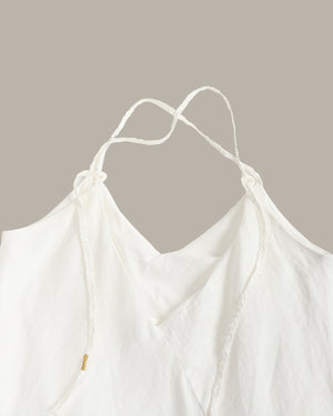 Close-up of a white camisole top with thin adjustable tie straps and a soft draped neckline, shown against a plain warm taupe background.