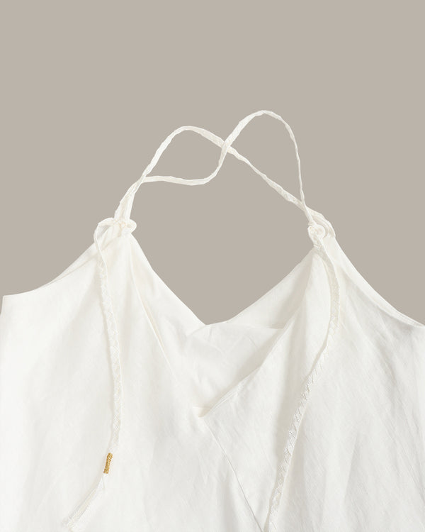Close-up of a white camisole top with thin adjustable tie straps and a soft draped neckline, shown against a plain warm taupe background.