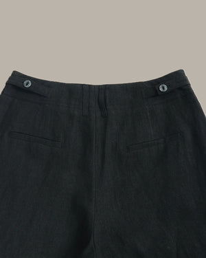 Close-up of the waistband and upper front of dark charcoal gray trousers, showing belt loops with metal buttons, front fly, and side welt pockets against a plain warm beige background.