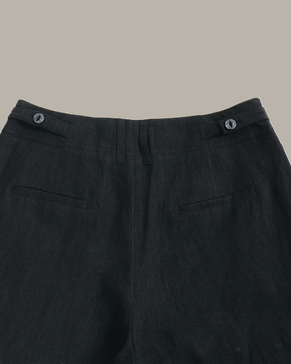 Close-up of the waistband and upper front of dark charcoal gray trousers, showing belt loops with metal buttons, front fly, and side welt pockets against a plain warm beige background.