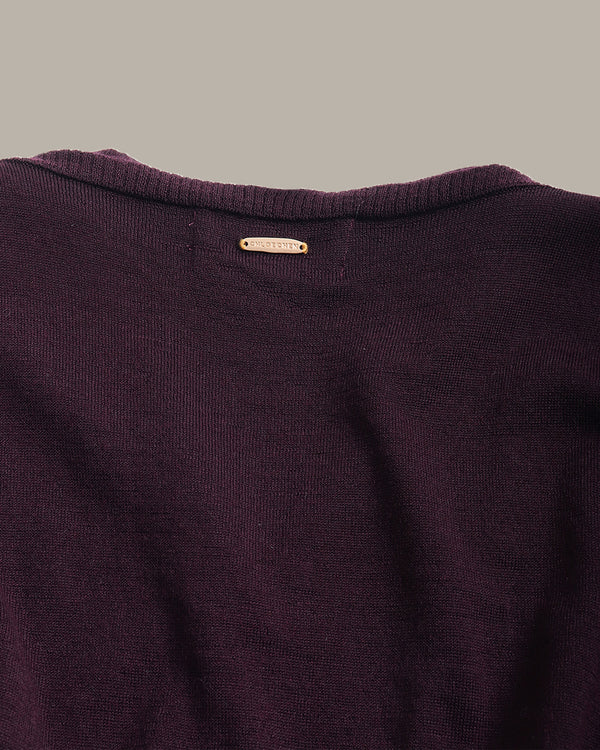 Close-up of a dark plum purple knit sweater showing the ribbed crew neck and a small gold-tone brand label that reads 'BURGA'.
