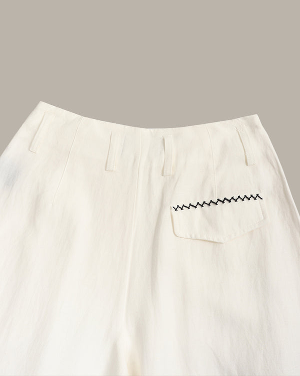 Close-up of off-white trousers showing the waistband, front fly, and a side pocket with black zigzag embroidery, set against a solid taupe background.