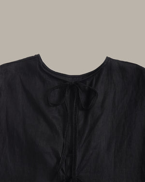 Close-up of a black top with a round neckline and a matching black ribbon tie at the front, shown against a solid taupe background.