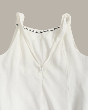 Close-up of a white sleeveless top with a V-neckline and delicate black embroidered detailing along the inner neckline, set against a solid taupe background.
