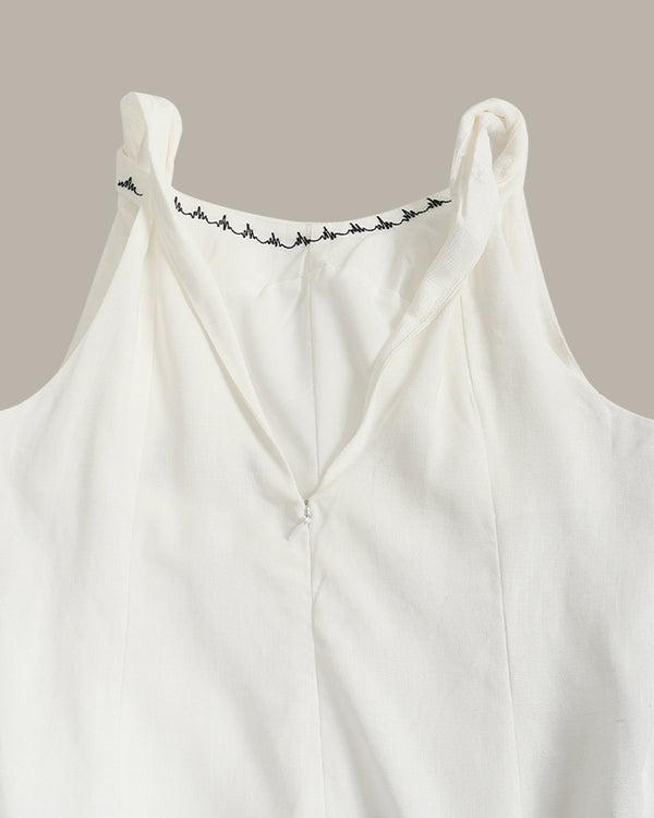 Close-up of a white sleeveless top with a V-neckline and delicate black embroidered detailing along the inner neckline, set against a solid taupe background.